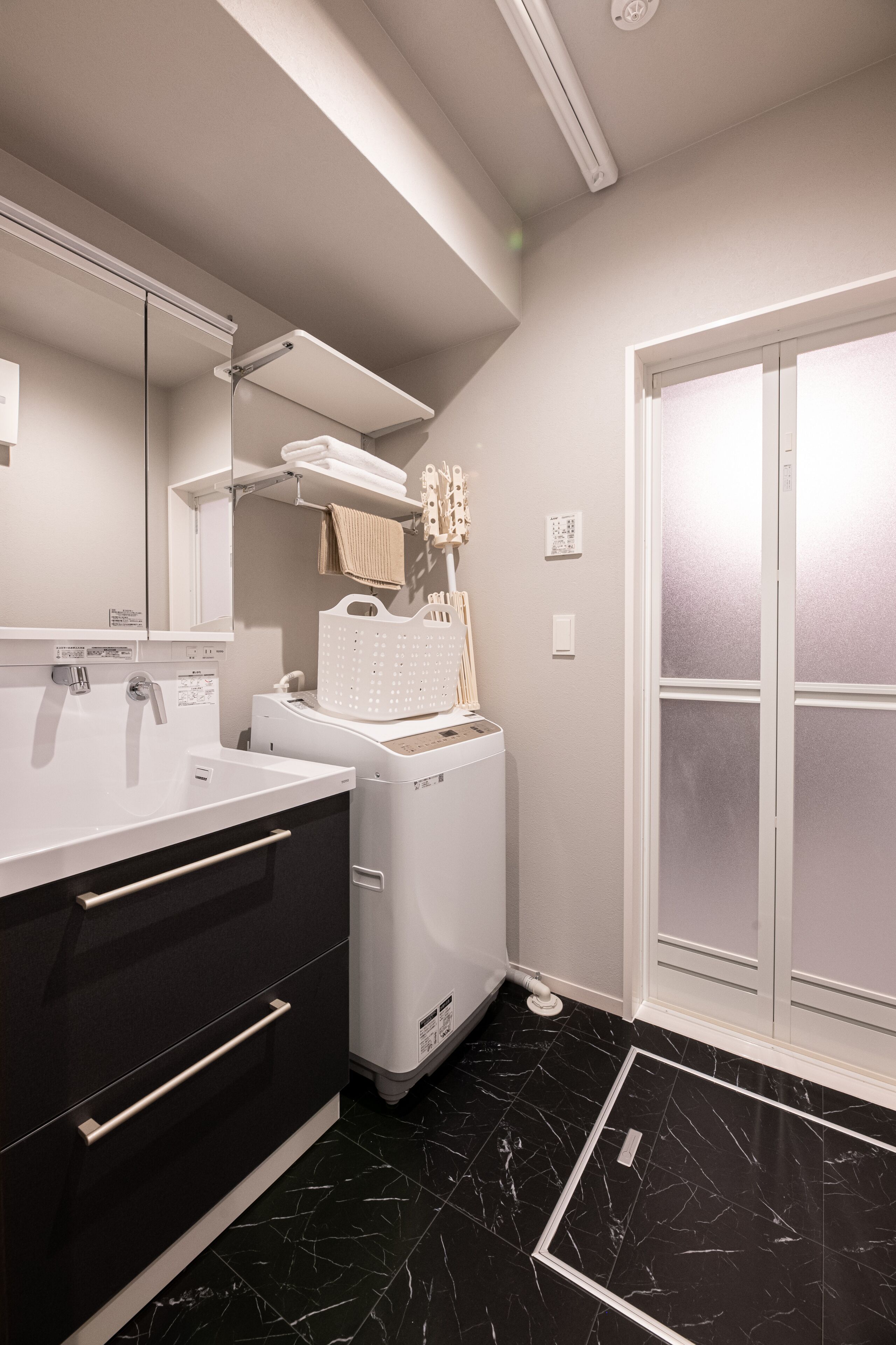 Un baño moderno con un espejo grande, un lavamanos blanco, suelo de mármol negro y una lavadora blanca.