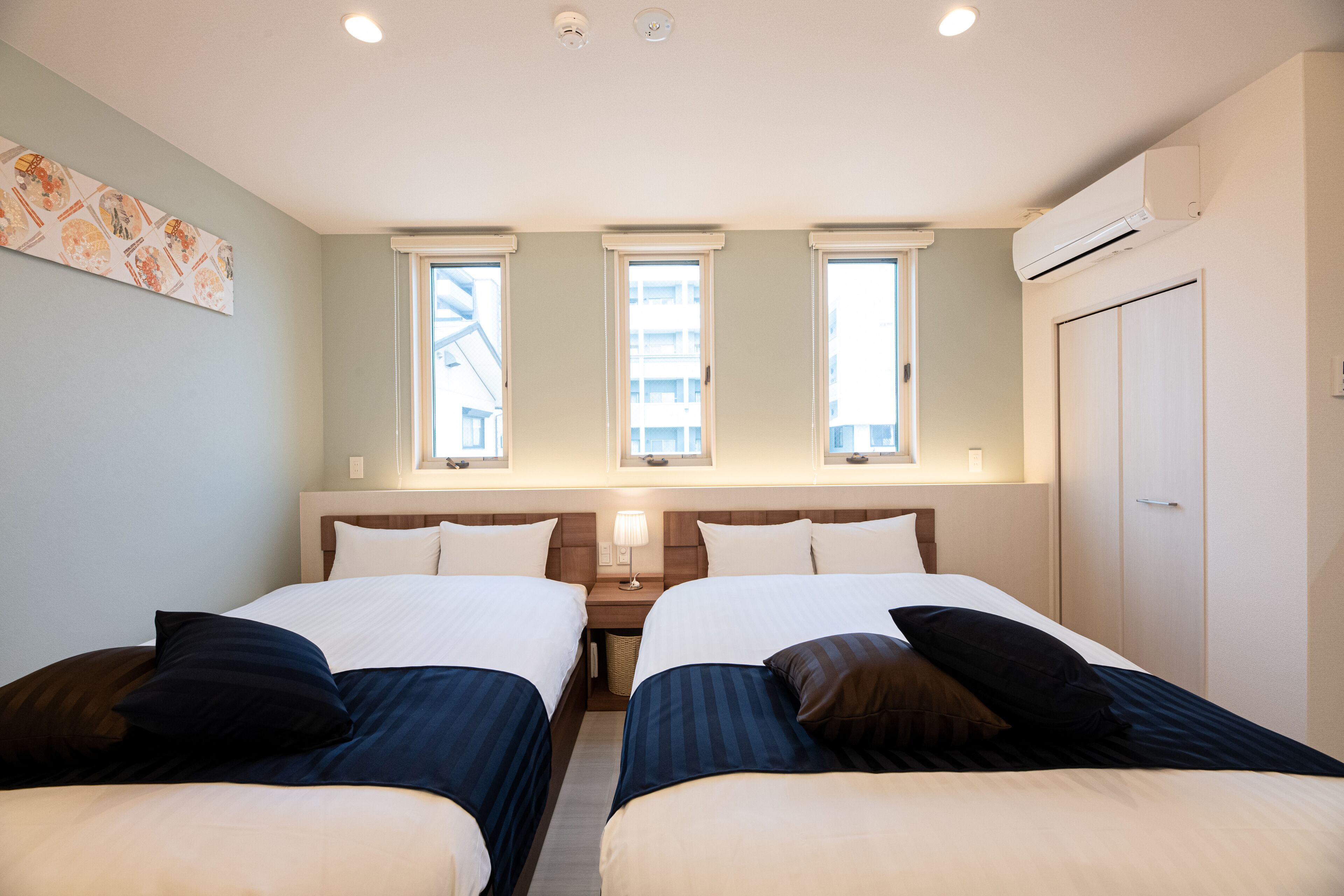 Habitación de hotel con dos camas, una mesita de noche, una lámpara y una ventana con vista a edificios.