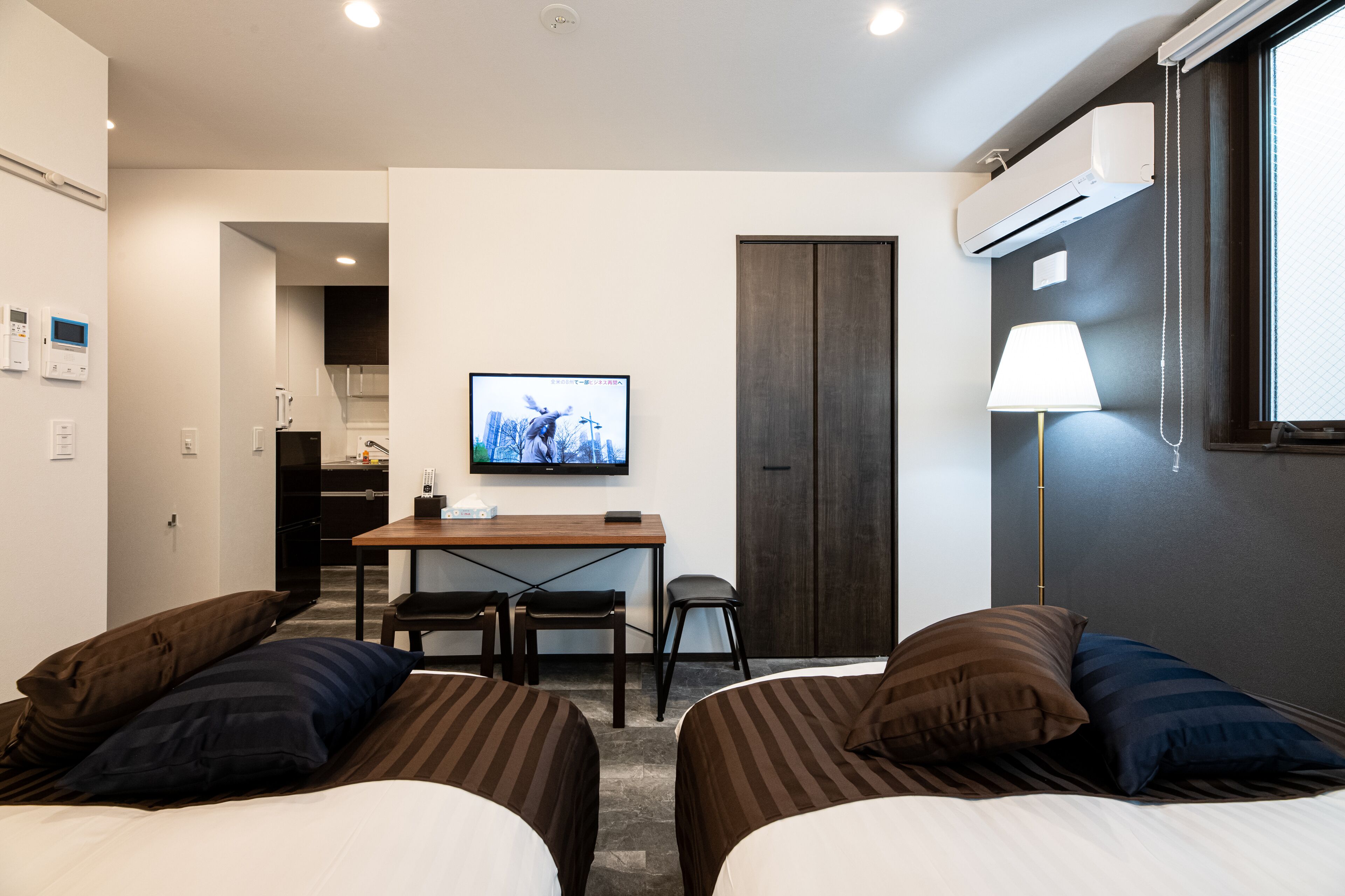 A hotel room with two beds, a desk with a chair, a television, an air conditioning unit, and a lamp.