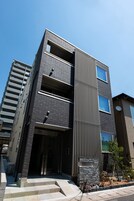 Un edificio moderno de varios pisos con un diseño vertical llamativo, una escalera que conduce a la entrada y un pequeño jardín con plantas.