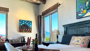 A hotel room with a bed, a tray with a bottle and wine glass, a painting, and a view of the mountains. - Secluded Desert Oasis: Luxurious Inn-Abiquiu Lake Views-Daily Gourmet Breakfast. (Abiquiu)