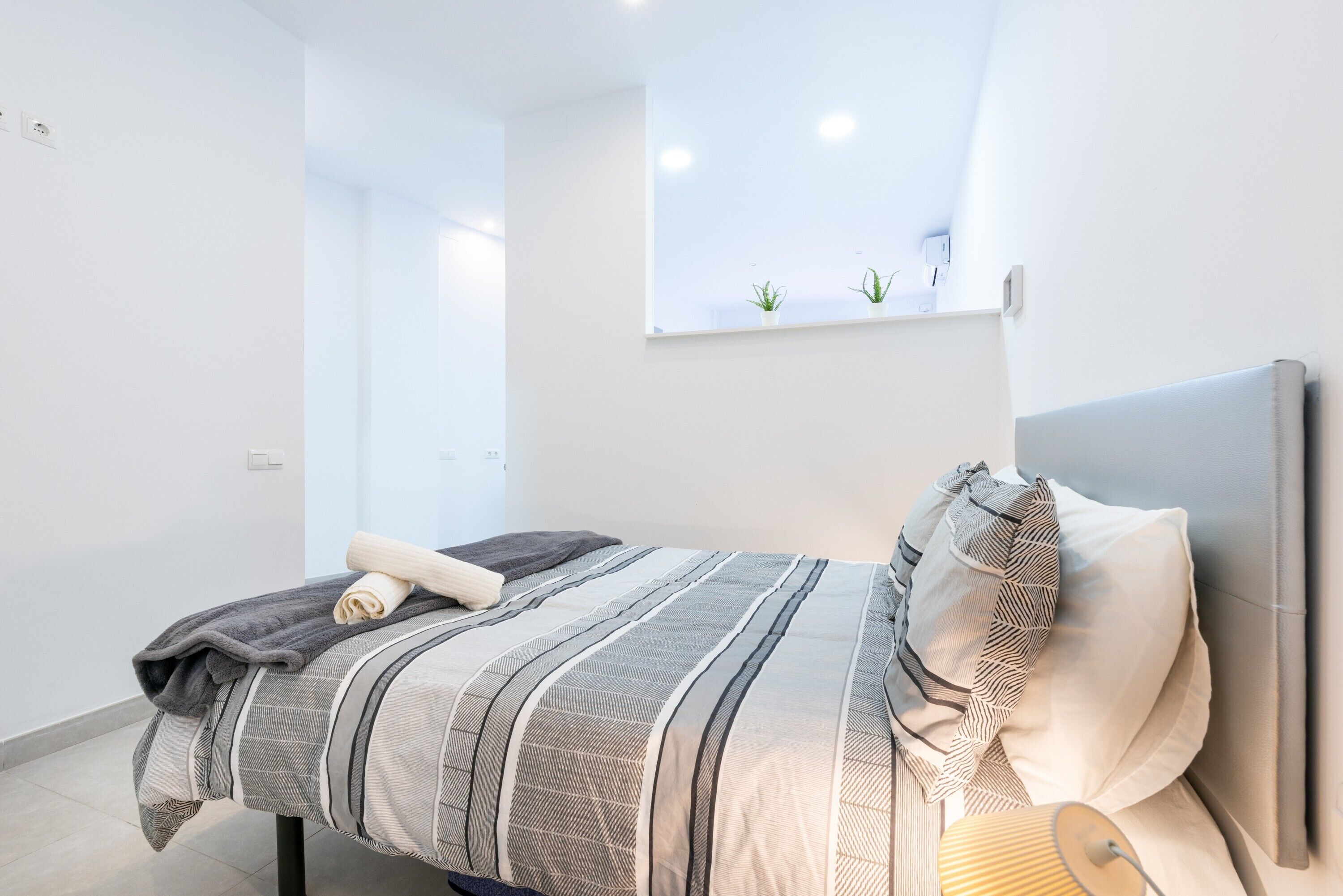 Ein modernes Schlafzimmer mit Bett, Nachttischlampe und einem kleinen Fenster mit Pflanzen.