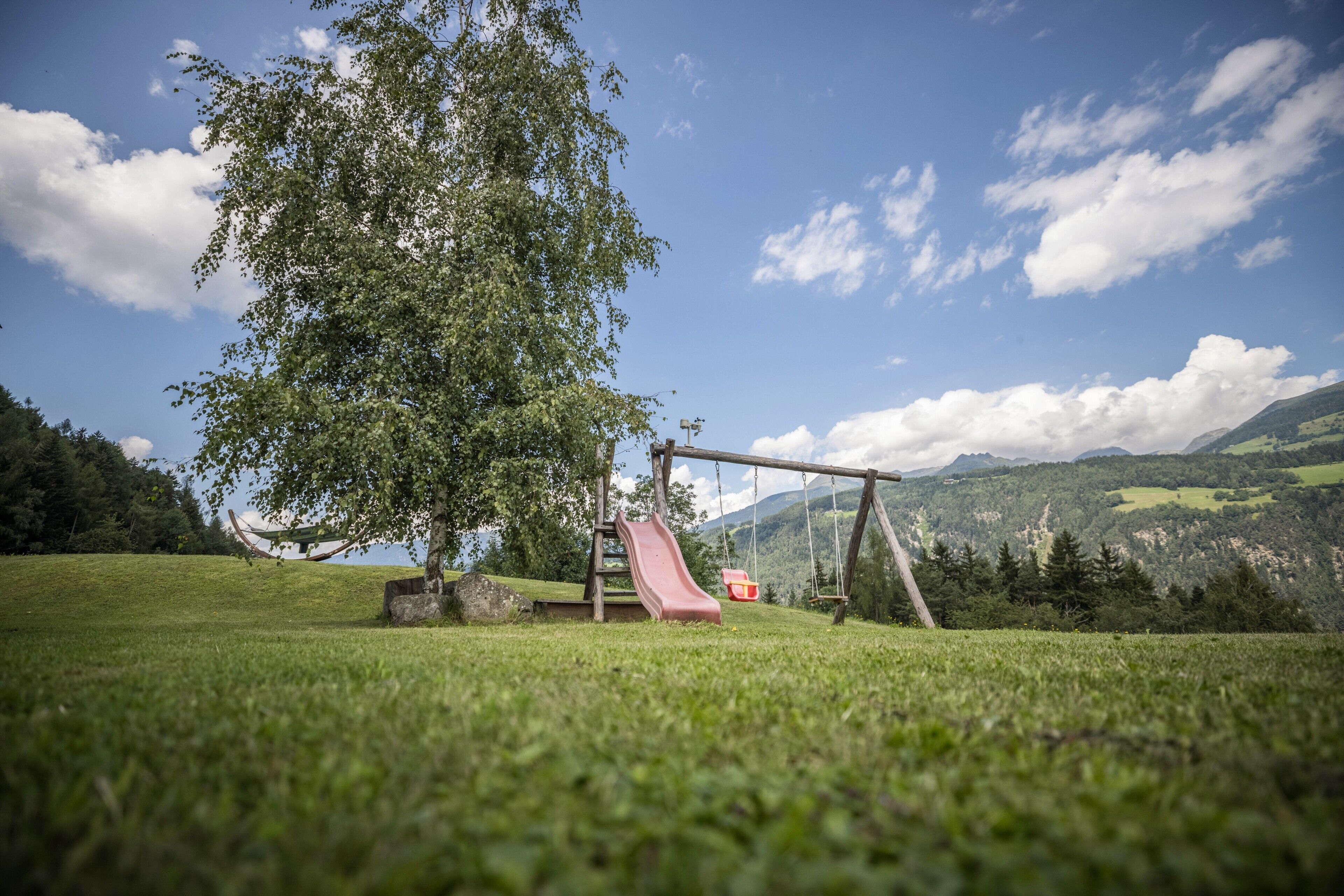 Un'area giochi con scivolo e altalene in un prato erboso, con montagne sullo sfondo.