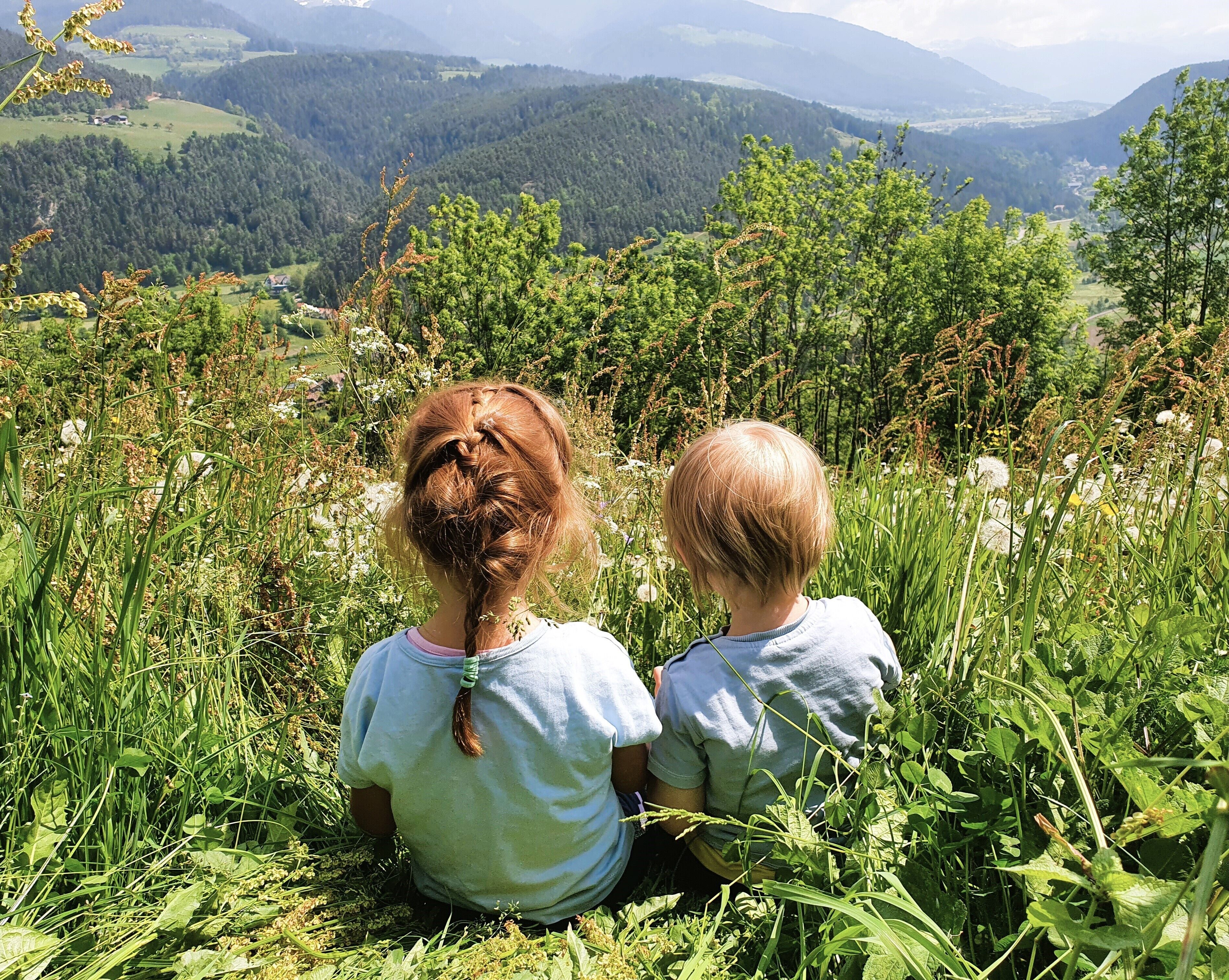 Twee kinderen zitten in een grasveld, met een bergachtig landschap op de achtergrond.
