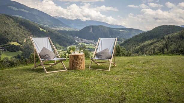 Deux chaises en bois, dotées de coussins, sont placées sur une colline herbeuse, avec une vue panoramique sur les montagnes en arrière-plan.