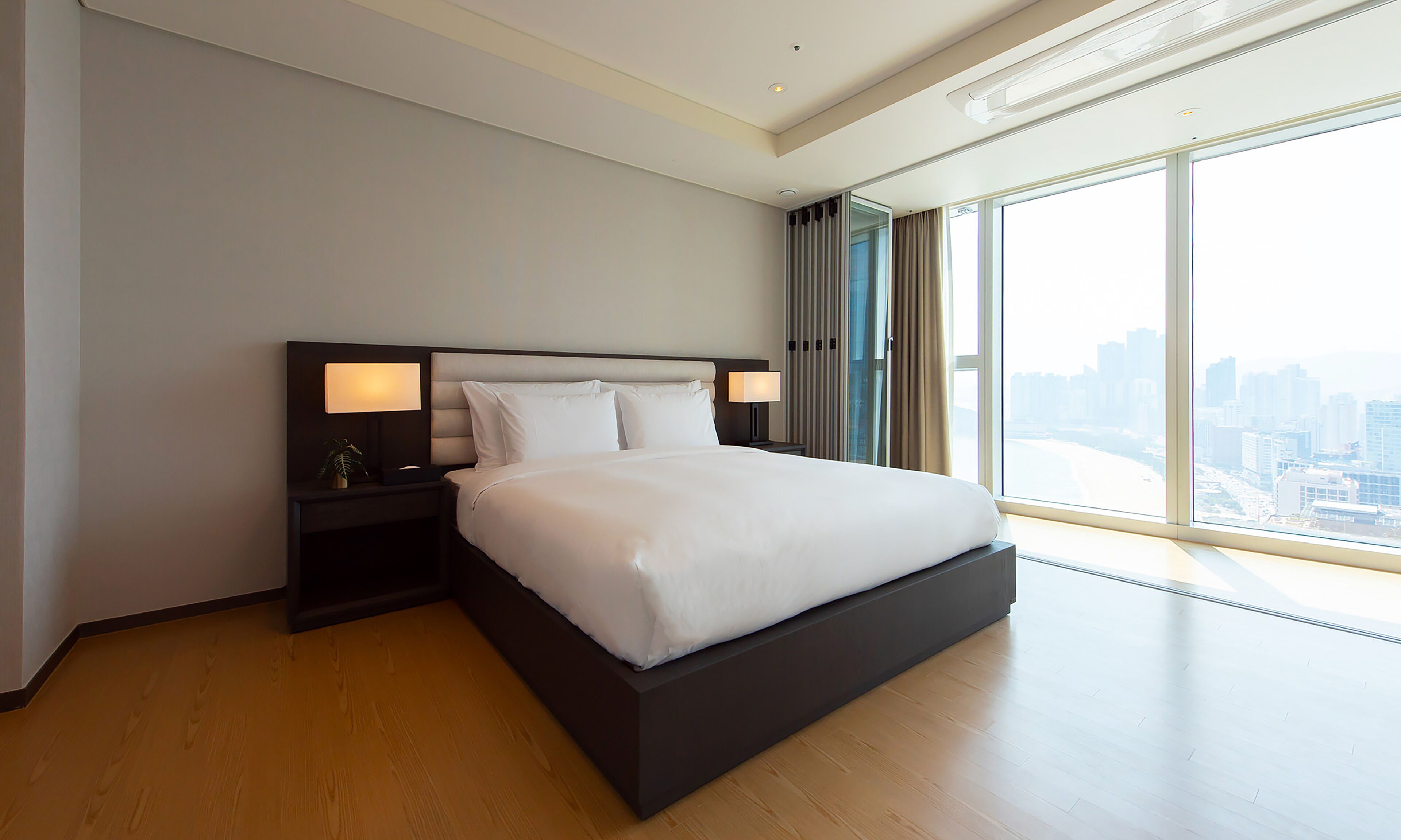 Quarto de hotel moderno com uma cama grande, duas mesas de cabeceira com luminárias, vista para a cidade através de janelas amplas e piso de madeira.