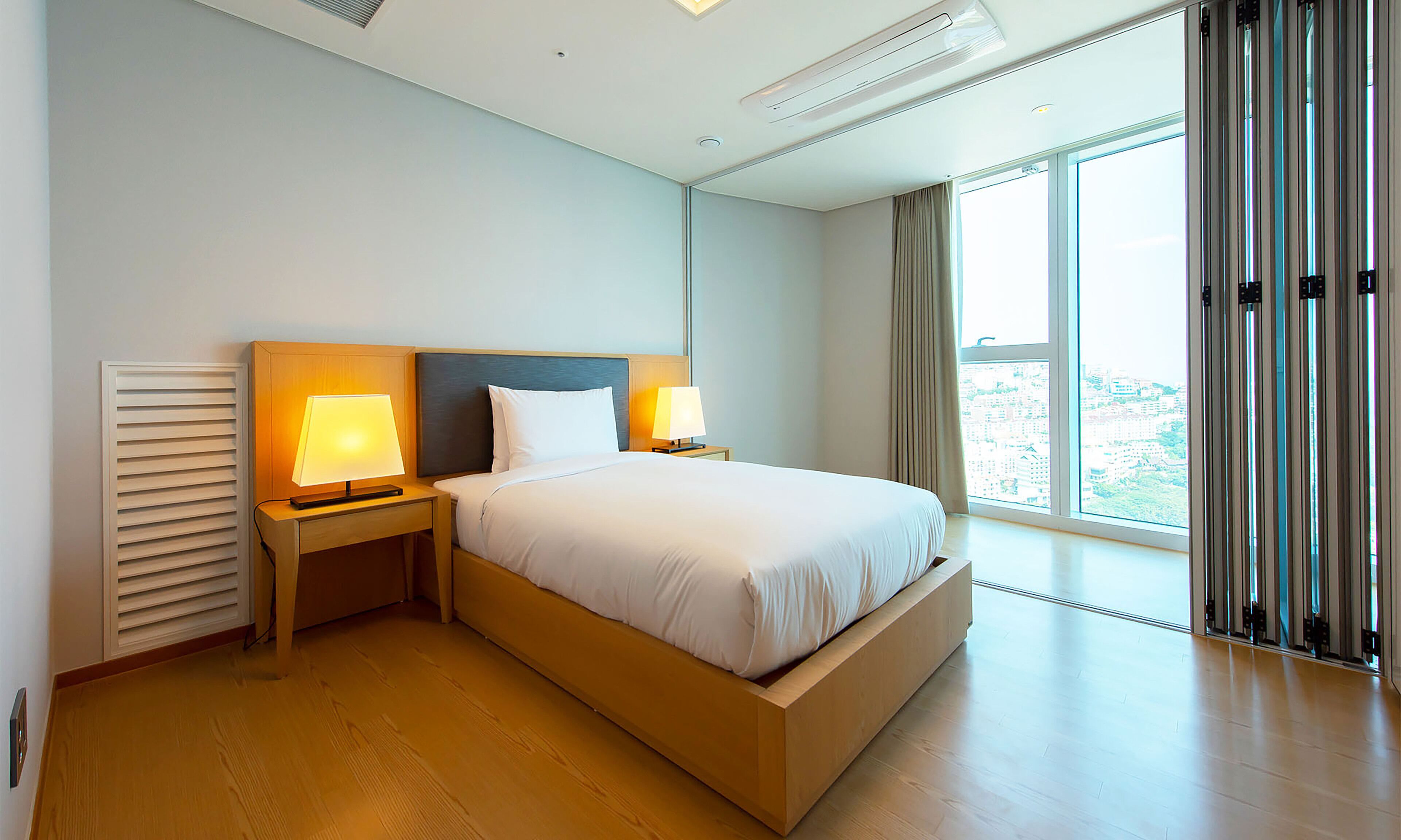 A modern hotel room with a large bed, two bedside lamps, a window with curtains, and a view of the city.