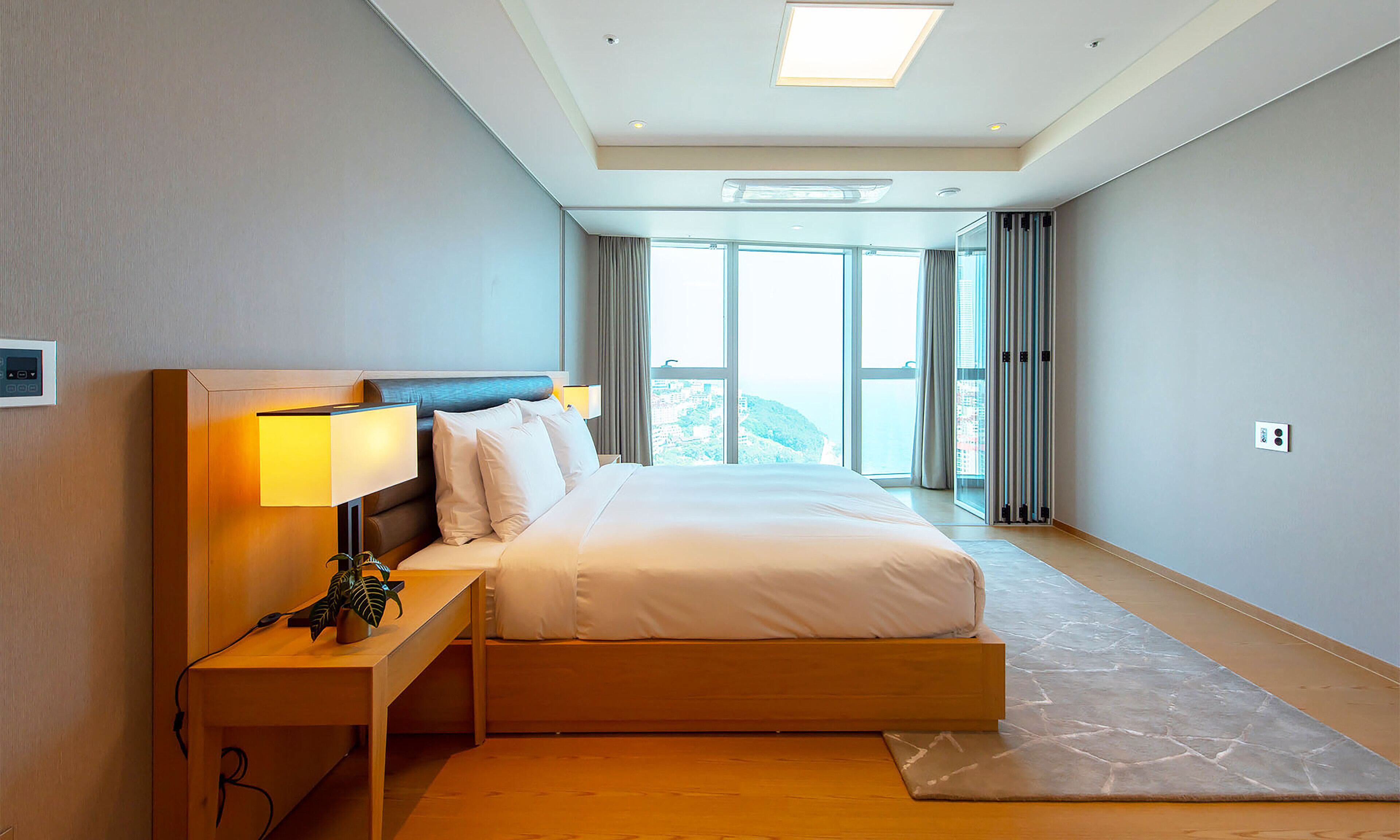 A modern hotel room with a large bed, a bedside table with a lamp, and a view of a cityscape through the window.
