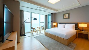 Una habitación de hotel moderna con una cama grande, un televisor de pantalla plana, un escritorio con lámpara y vistas a la ciudad a través de amplias ventanas.