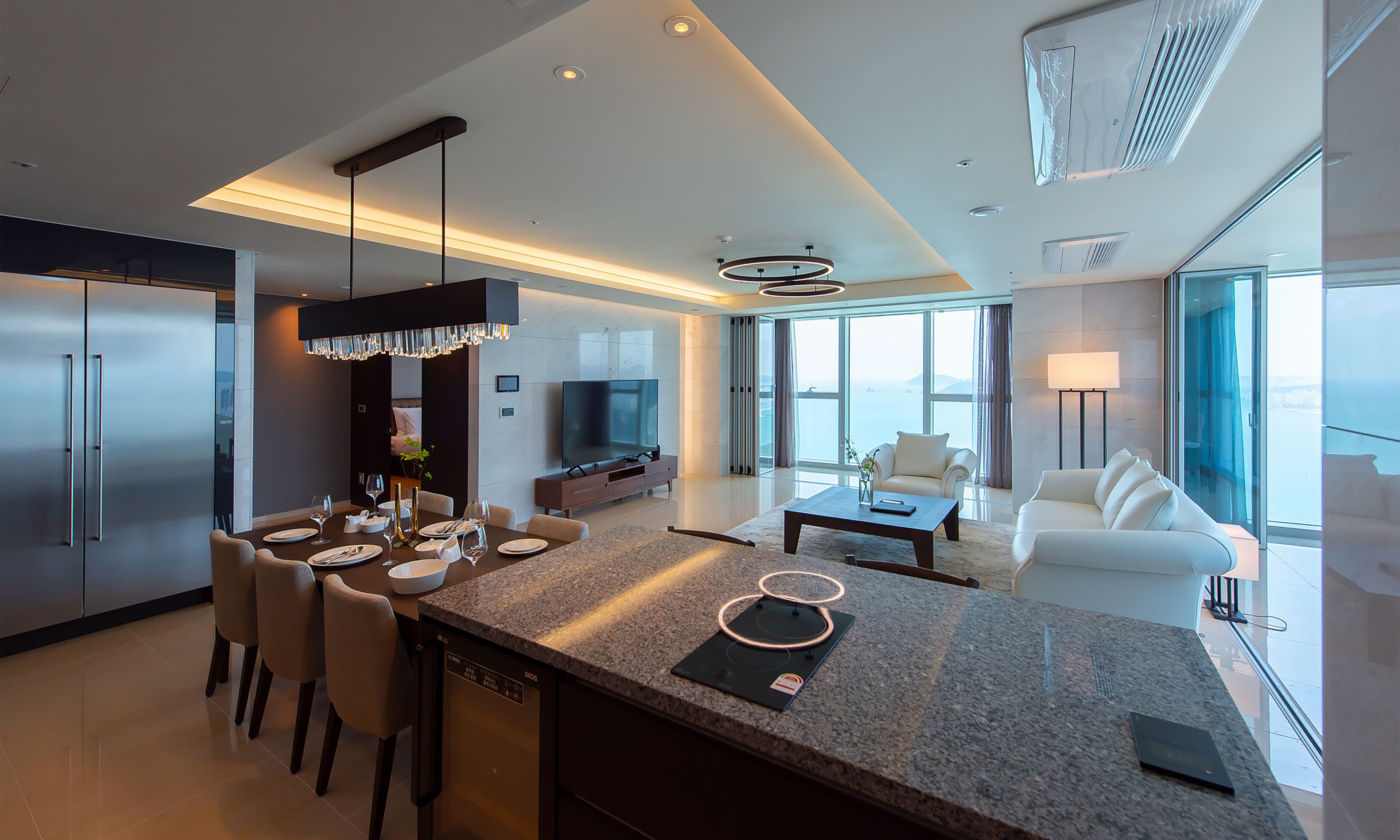 A modern hotel room with a kitchen, dining area, and living space. It features a large island with bar tools, a flat-screen TV, and a seating area with a sofa and armchairs.