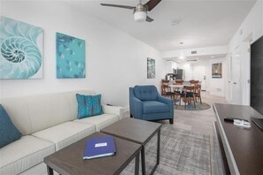 Un salon moderne avec un canapé, une table basse et un fauteuil bleu. On aperçoit à l’arrière-plan un coin repas avec une table et des chaises.