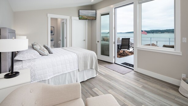 A bedroom with a large bed, a desk, and a chair. There is a sliding glass door leading to a balcony with a view of the water and an American flag. - AC, kayaks, rooftop deck with fire pit, boat launch and incredible views! (Camano)