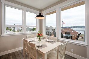 Un comedor con una mesa dispuesta para cuatro personas, ventanas amplias que dan vista a una zona residencial y una masa de agua.