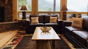 A cozy living room with a fireplace, two leather sofas with moose pillows, a wooden coffee table, and large windows offering a view of mountains.
