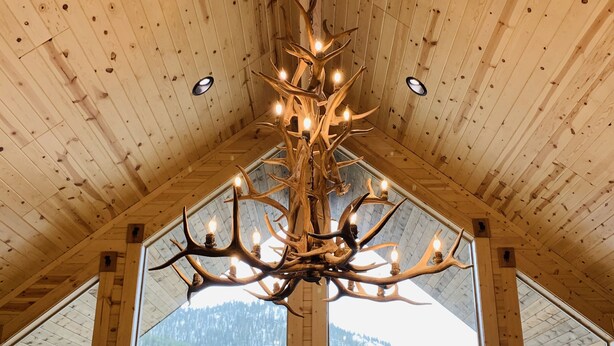 A wooden interior with a chandelier made of antlers, large windows, and a view of a snowy landscape.