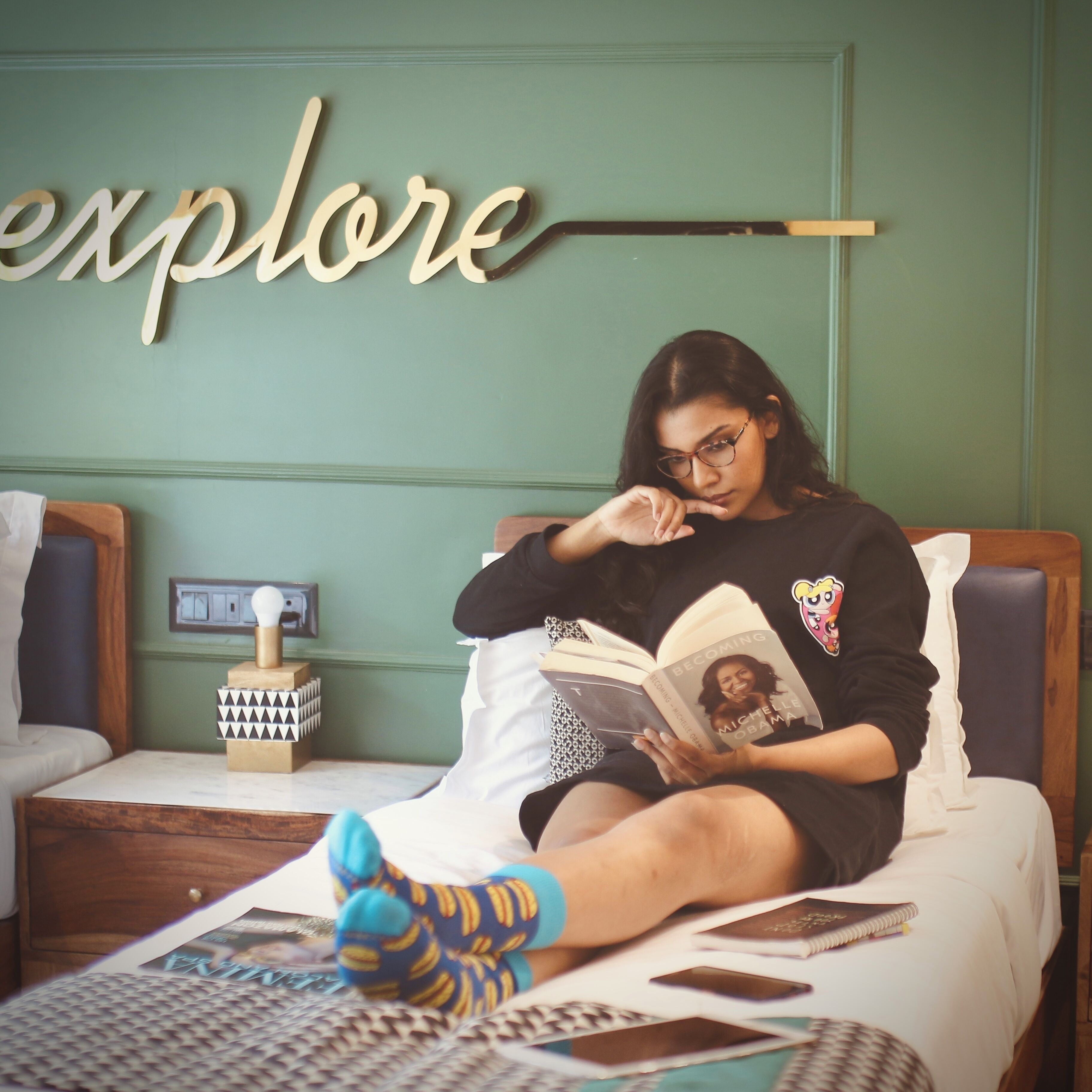 Una persona sentada en una cama leyendo un libro en una habitación con una pared verde y un letrero que dice 'explore'.