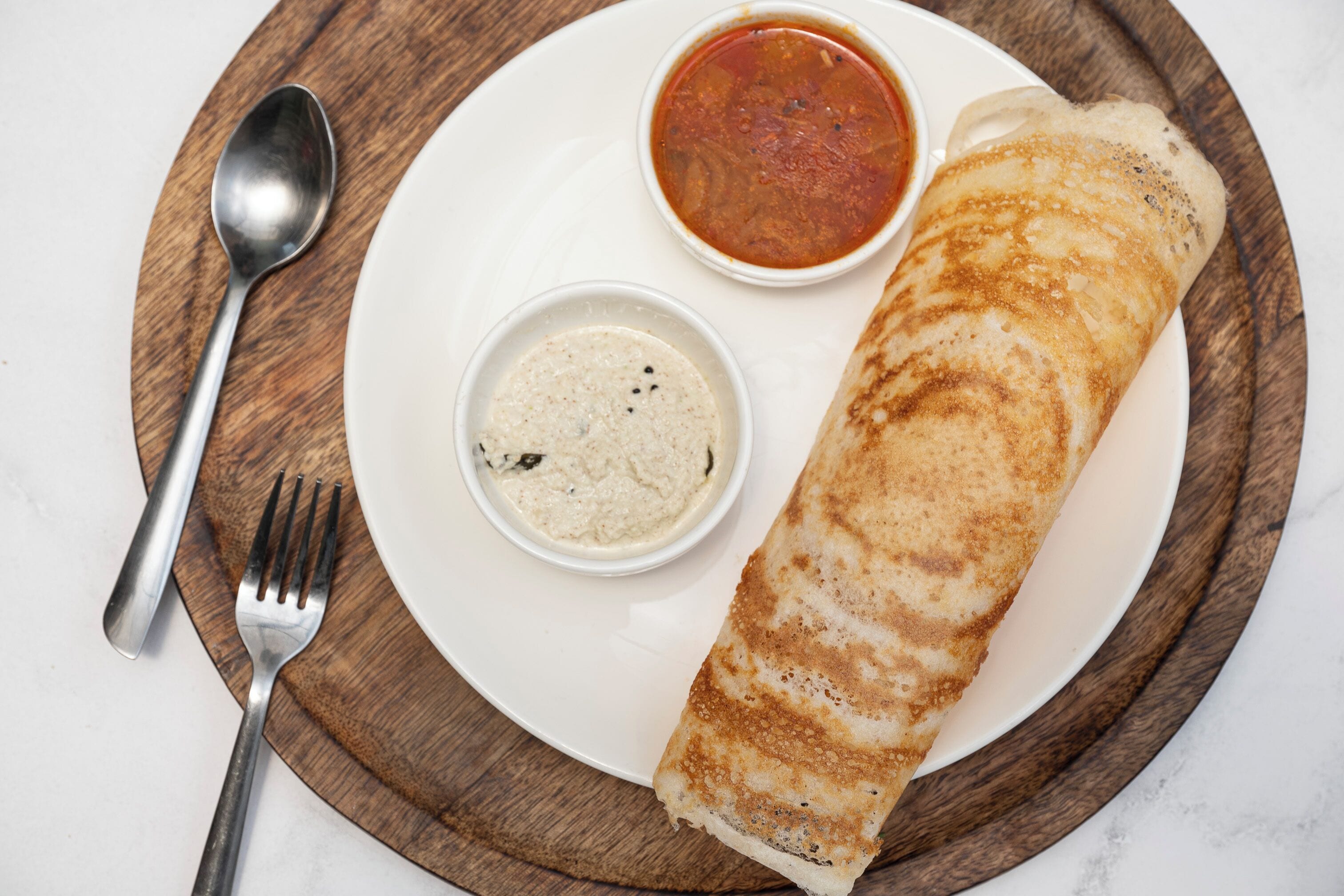 A plate with a rolled flatbread, two small bowls of dips, a spoon, and a fork on a wooden board.