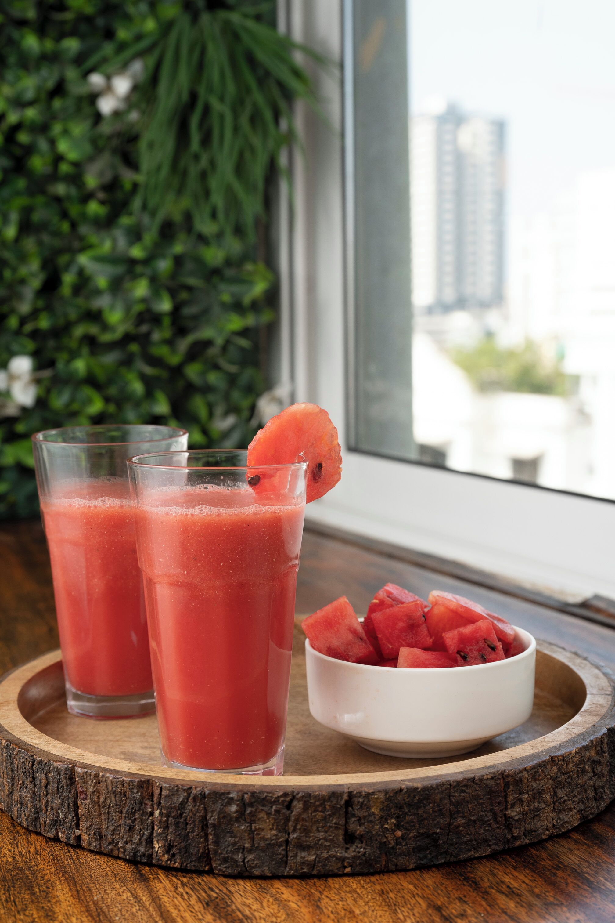 Two glasses of pink beverage on a wooden tray with a bowl of watermelon slices beside them.