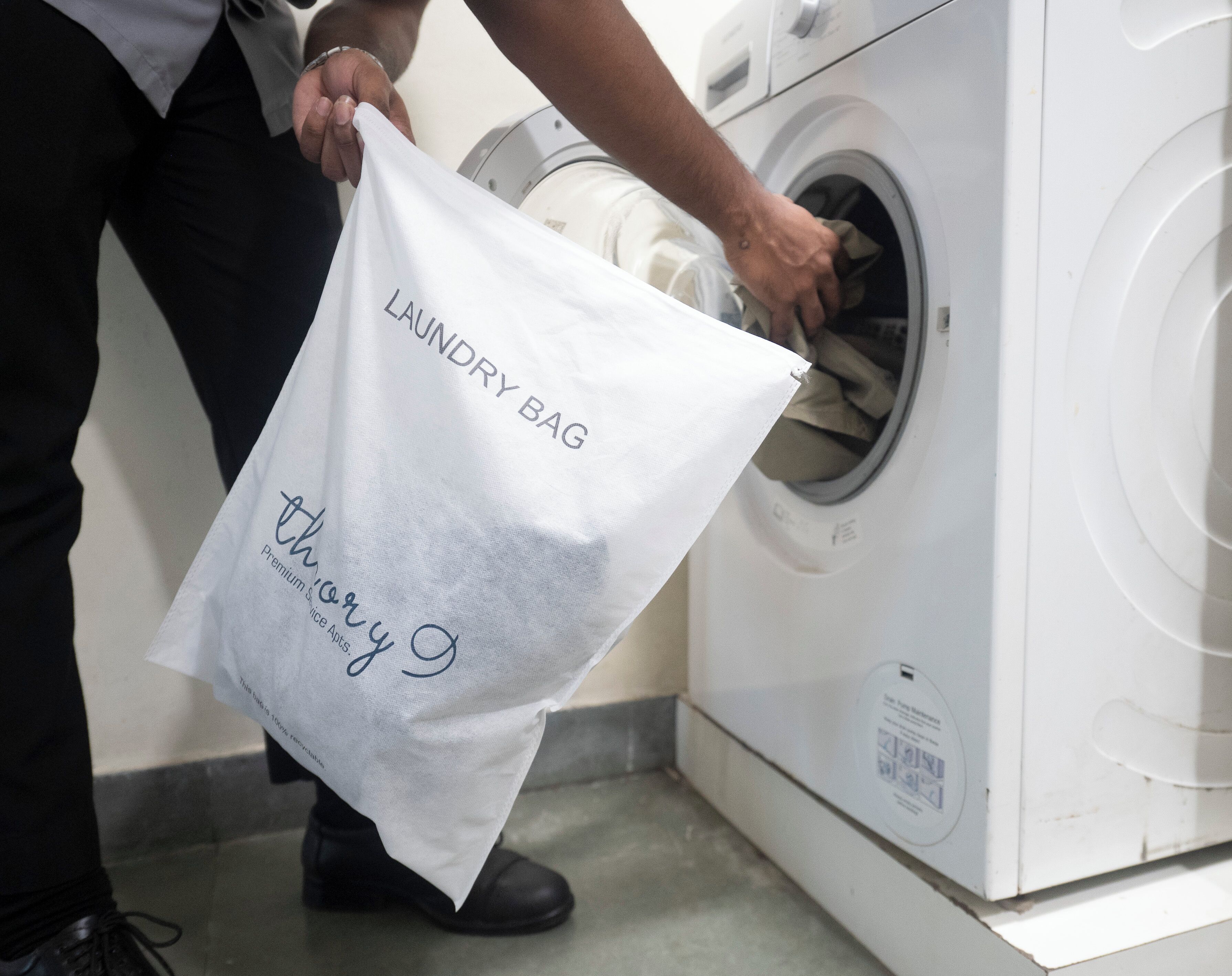 Een persoon houdt een witte waszak met tekst vast, staand naast een wasmachine.