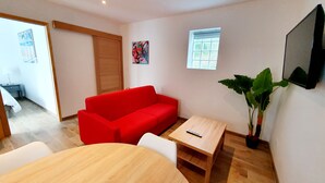 Ein gemütlicher Wohnraum mit einem roten Sofa, einem Holztisch, einem an der Wand befestigten Fernseher und einem Blumentopf.