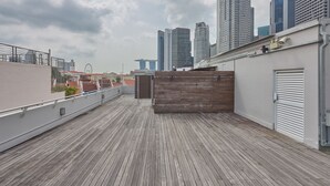 Terraço no telhado com deck de madeira, parede branca e vista para edifícios altos ao fundo.