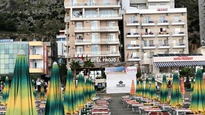 Un hotel llamado FROJD con varios pisos y balcones, una zona de playa con tumbonas y sombrillas, y montañas al fondo.