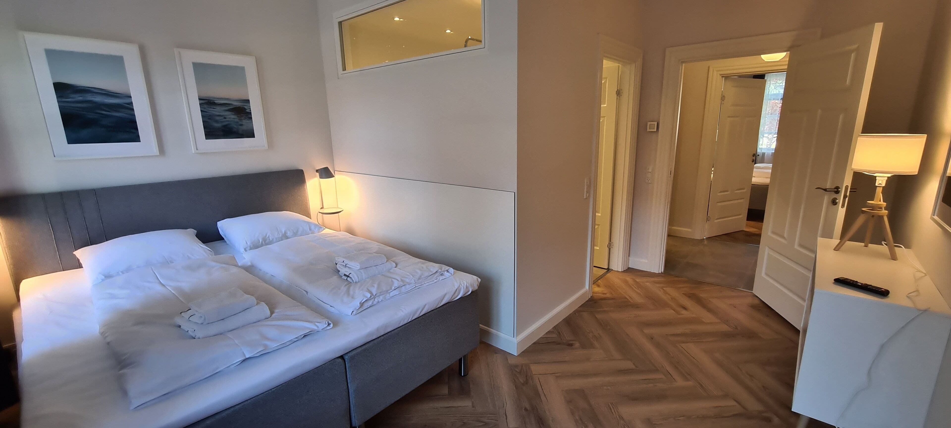 Una cama bien hecha con almohadas y toallas dobladas, una lámpara de noche y un espejo montado en la pared.