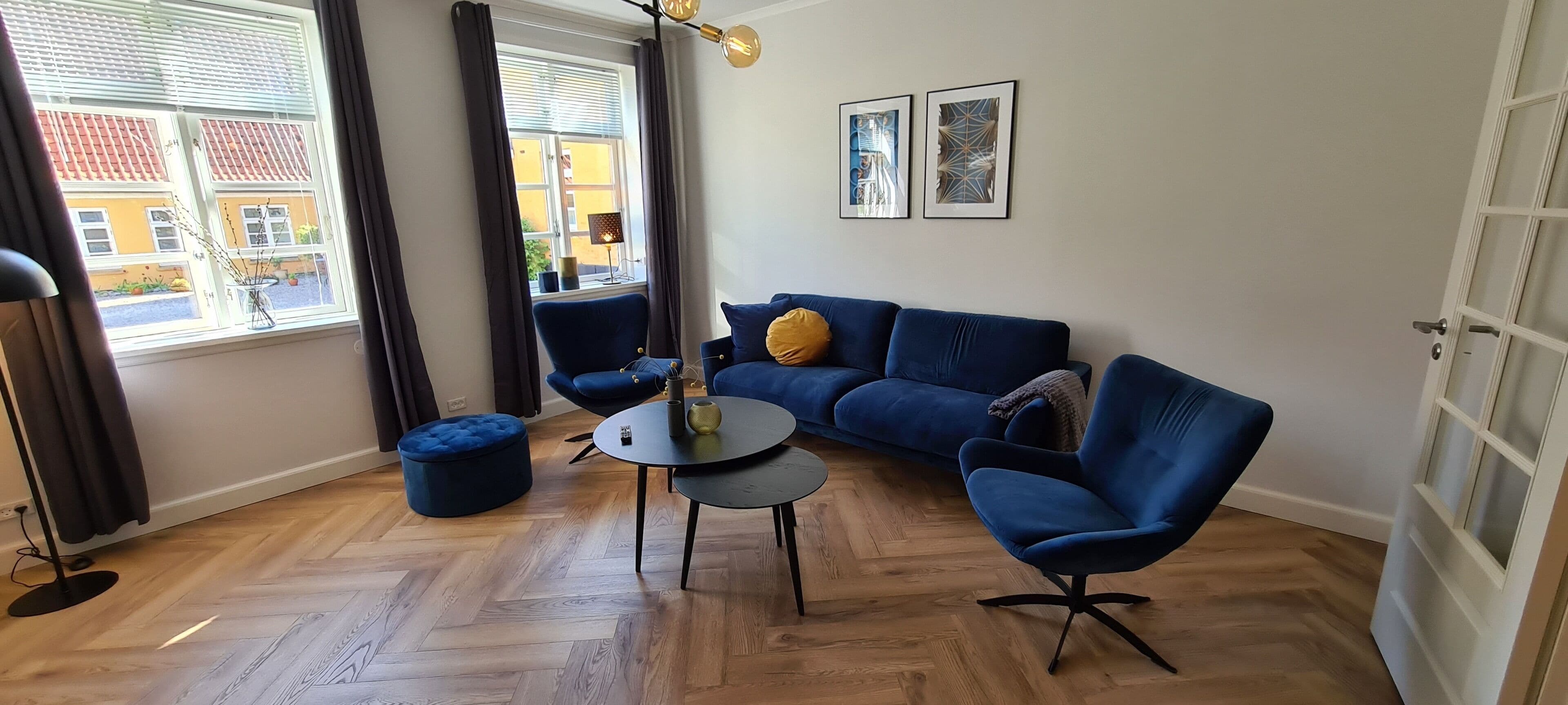 A living room with a blue sofa, a round coffee table, and a blue armchair. There is a window with curtains, a lamp, and two framed pictures on the wall.