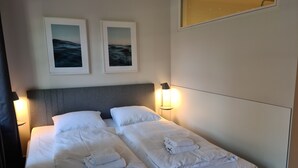 Una cama bien tendida con sábanas y toallas blancas, un cabecero gris y dos cuadros en la pared.