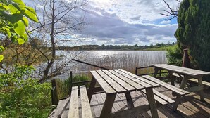 Terraza de madera con mesas de picnic dando a un lago, rodeada de follaje y árboles.