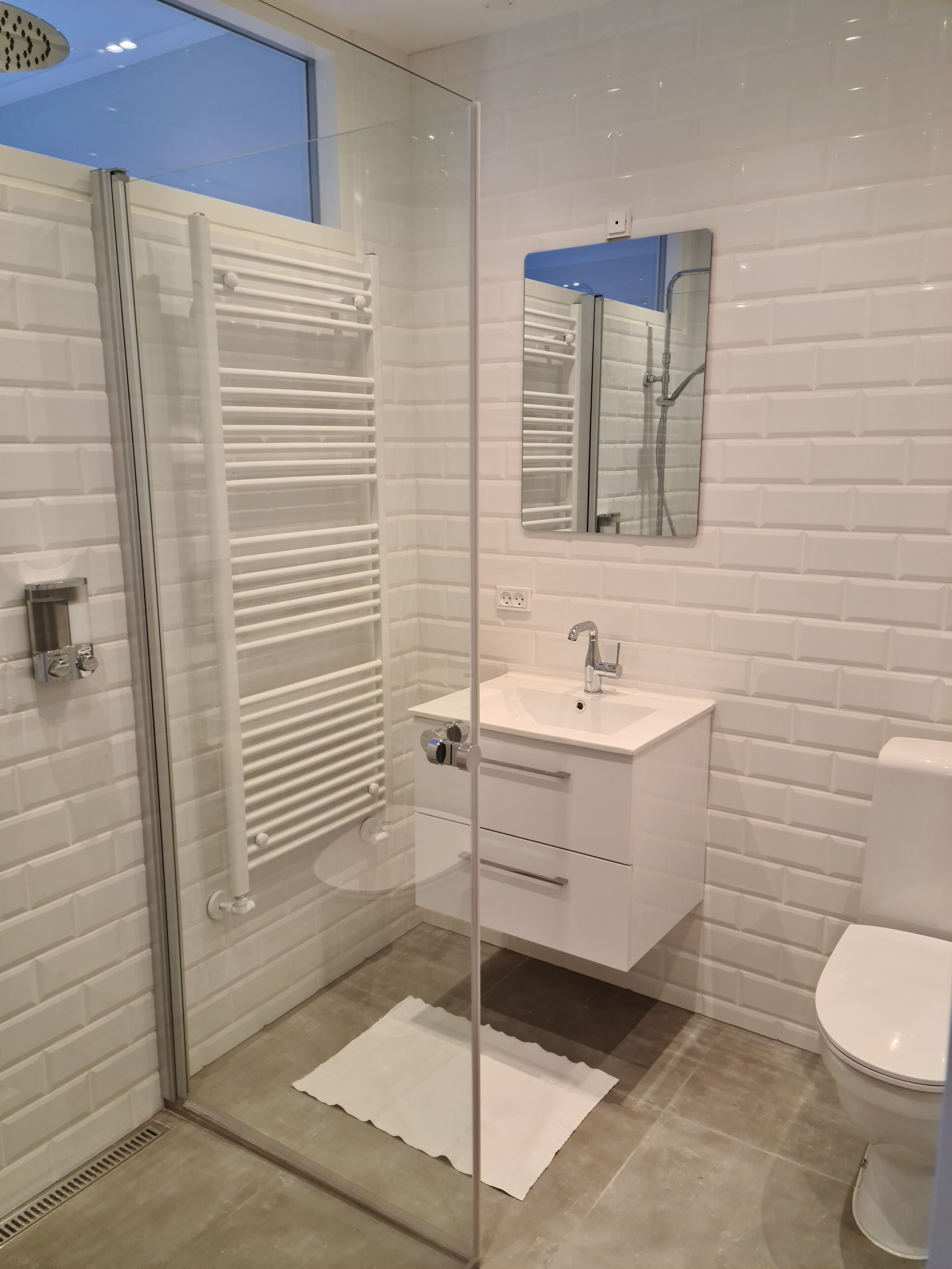 Une salle de bain moderne avec une douche à parois de verre, des murs recouverts de carreaux blancs, un lavabo blanc et des toilettes.