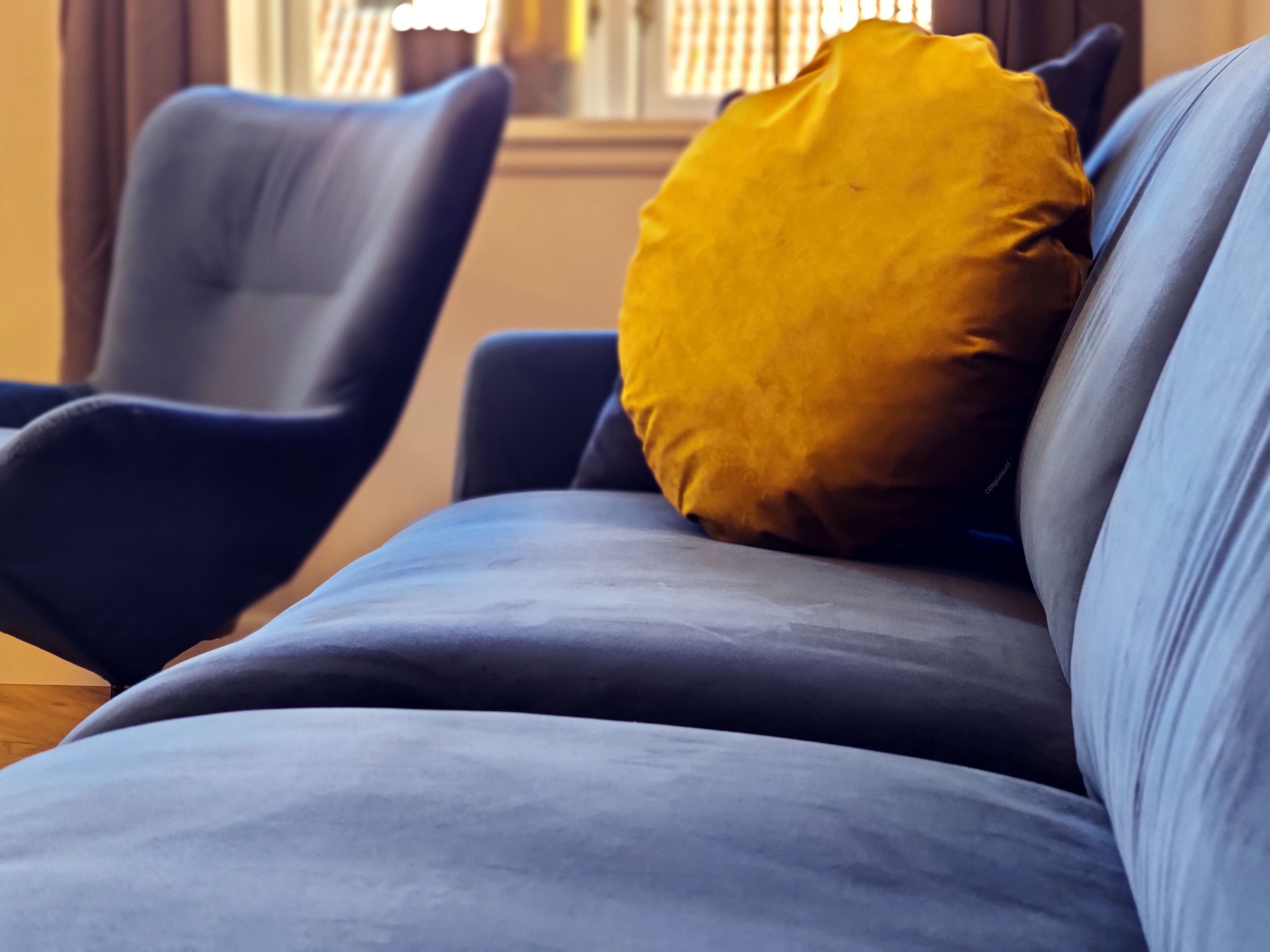 Un canapé bleu avec un coussin jaune et un fauteuil bleu foncé dans une pièce dont on aperçoit une fenêtre au fond.