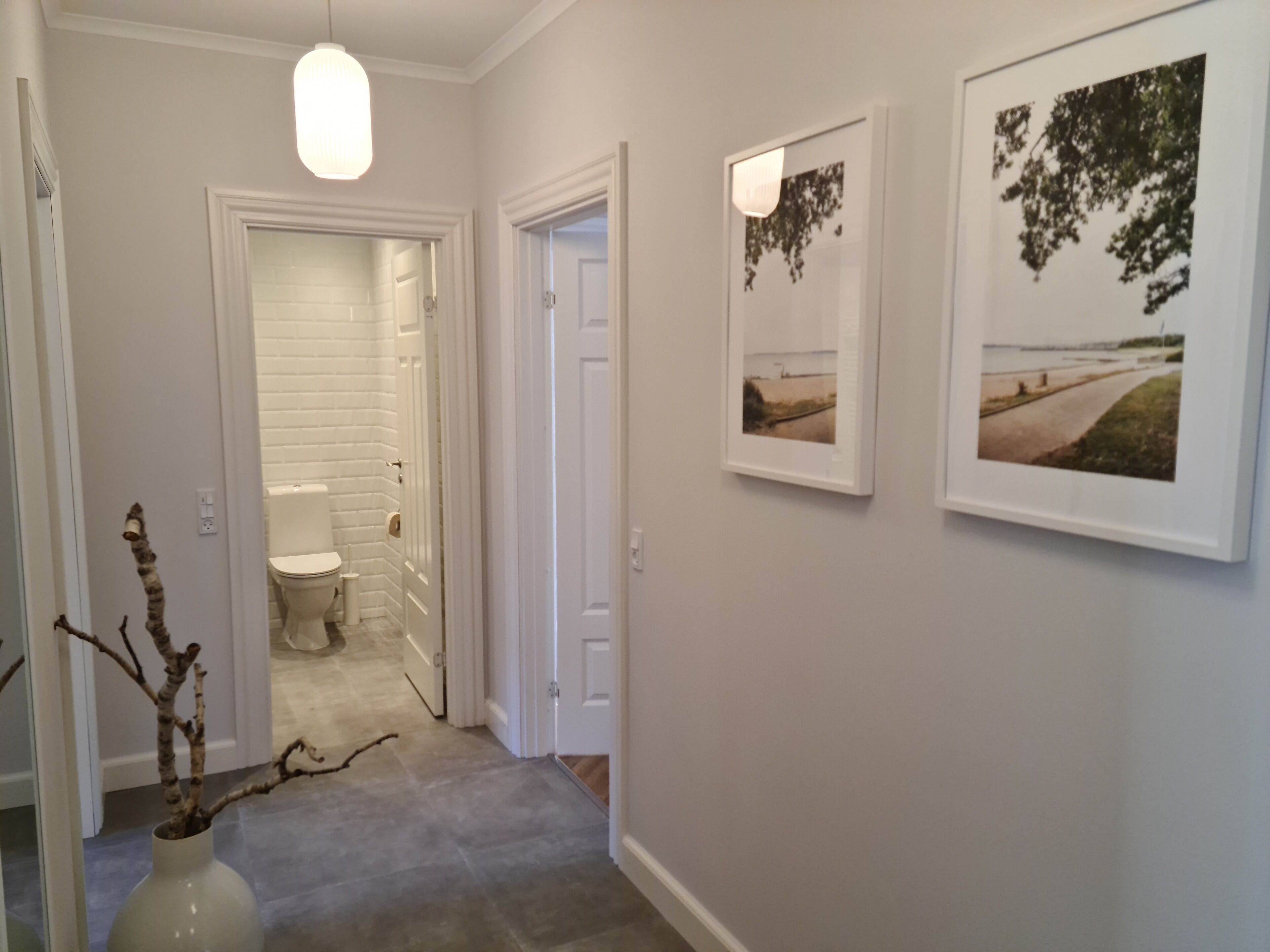 A hallway with a white door leading to a bathroom, a vase with branches, and two framed pictures on the wall.