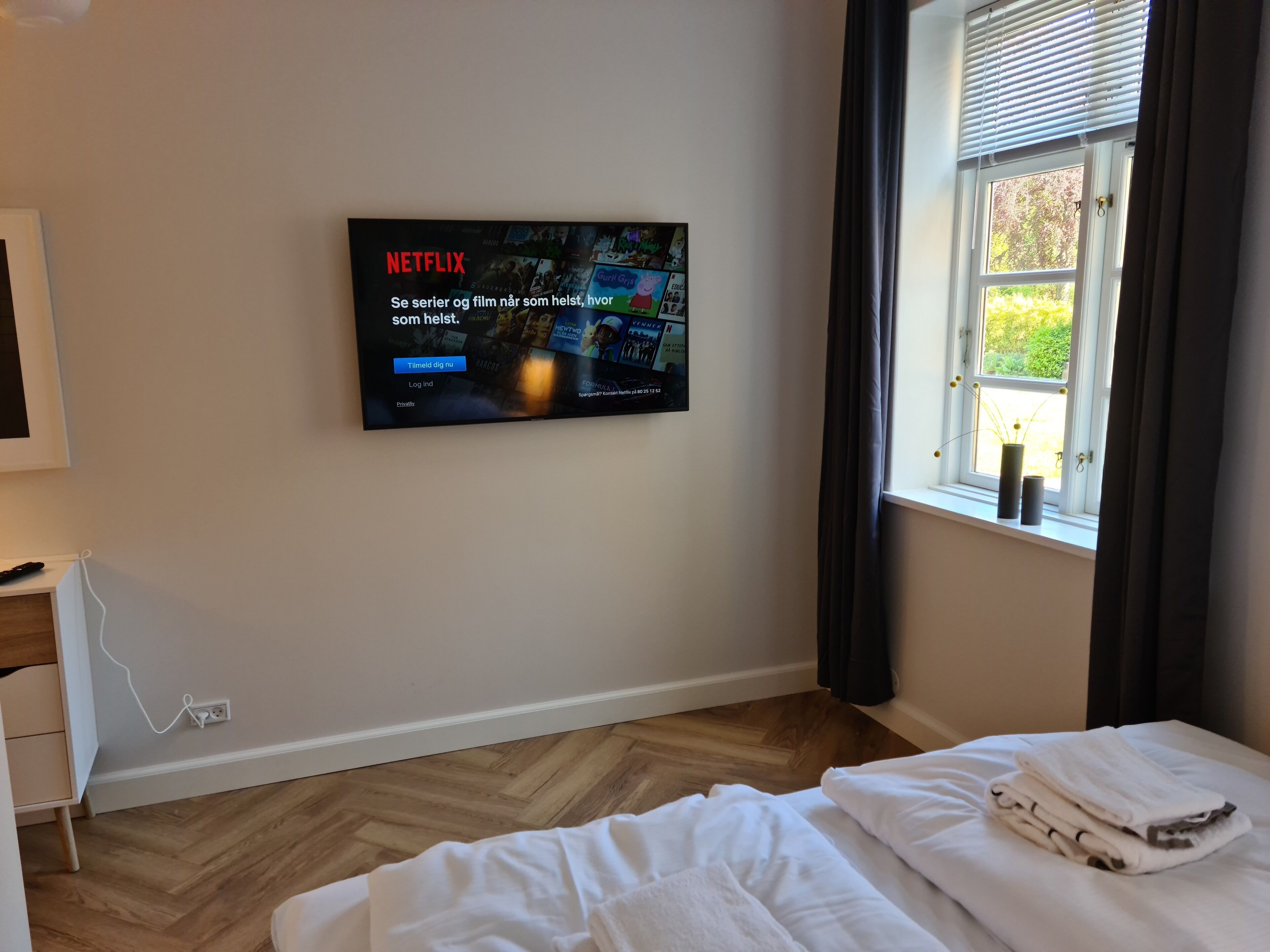 Ein Hotelzimmer mit einem großen Flachbildfernseher an der Wand, der das Netflix-Logo zeigt. Ein Bett mit weißen Leinentüchern und einem gefalteten Handtuch ist ebenfalls zu sehen. Ein Fenster mit dunklen Vorhängen ist erkennbar, und ein kleiner Tisch mit einer Fernbedienung befindet sich auf der linken Seite des Zimmers.