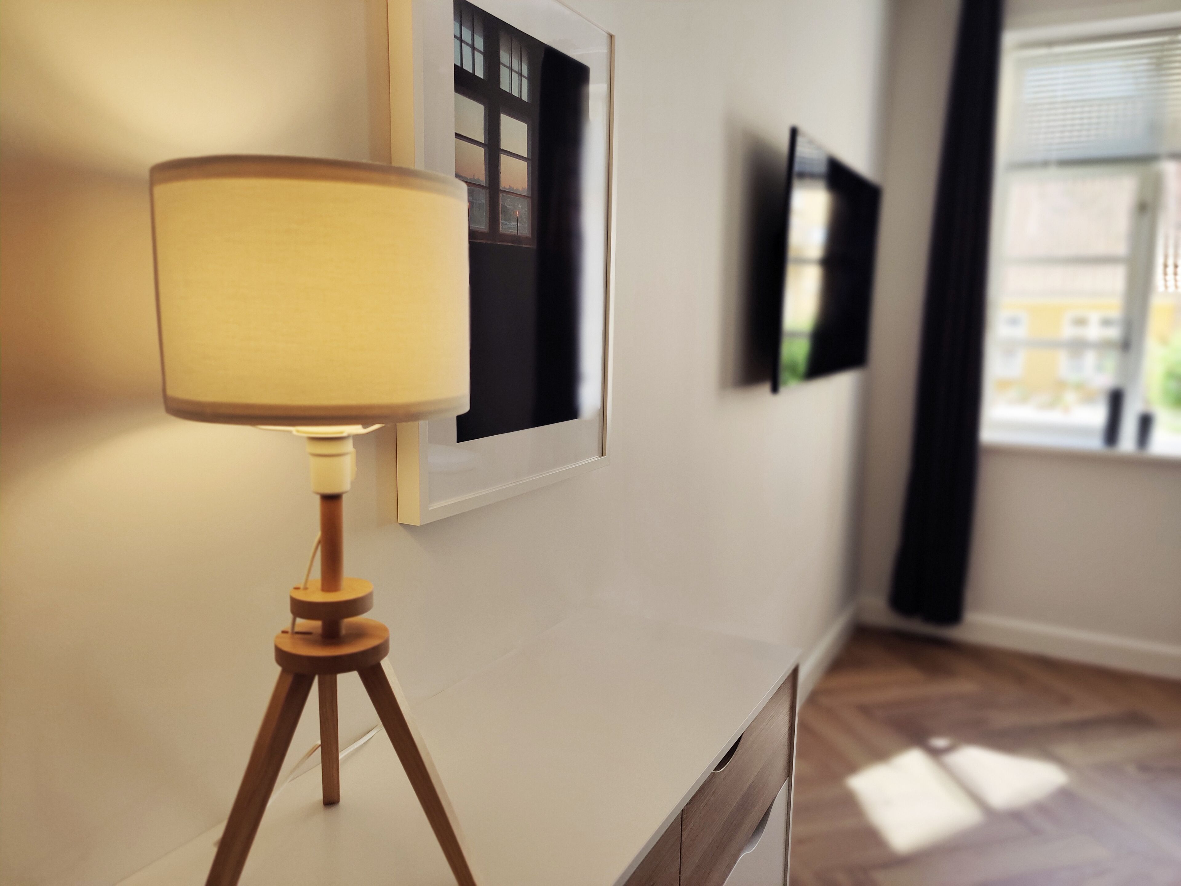 Een kamer met een vloerlamp op een statief, een lijst met een afbeelding aan de muur, en een raam met jaloezieën.