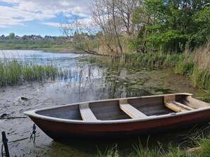 A small wooden boat tied to a dock by a calm lake with surrounding greenery and houses in the background. - The Old Mill - By the Lake (Krusaa)