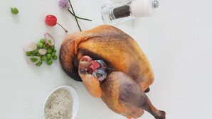 Een hele gebraden kip op een wit snijplank, met gesneden groene uien, een kleine schaal rijst, een pepermolen en een takje bloemen.