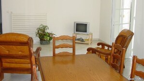 Een eetgedeelte met een houten tafel en stoelen, een televisie op een standaard, een potplant en een witte radiator.
