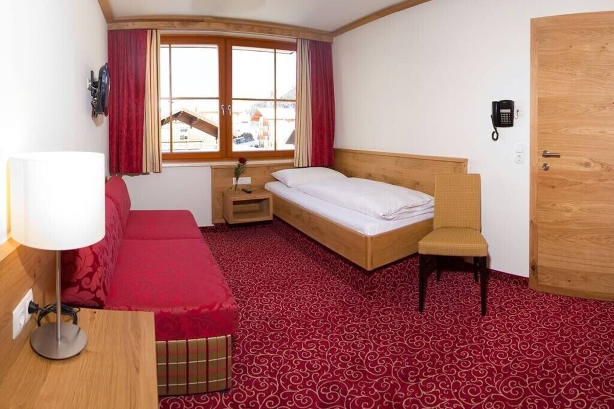 A hotel room with a single bed, a small table, a chair, a desk, a lamp, a telephone, and a window with curtains.