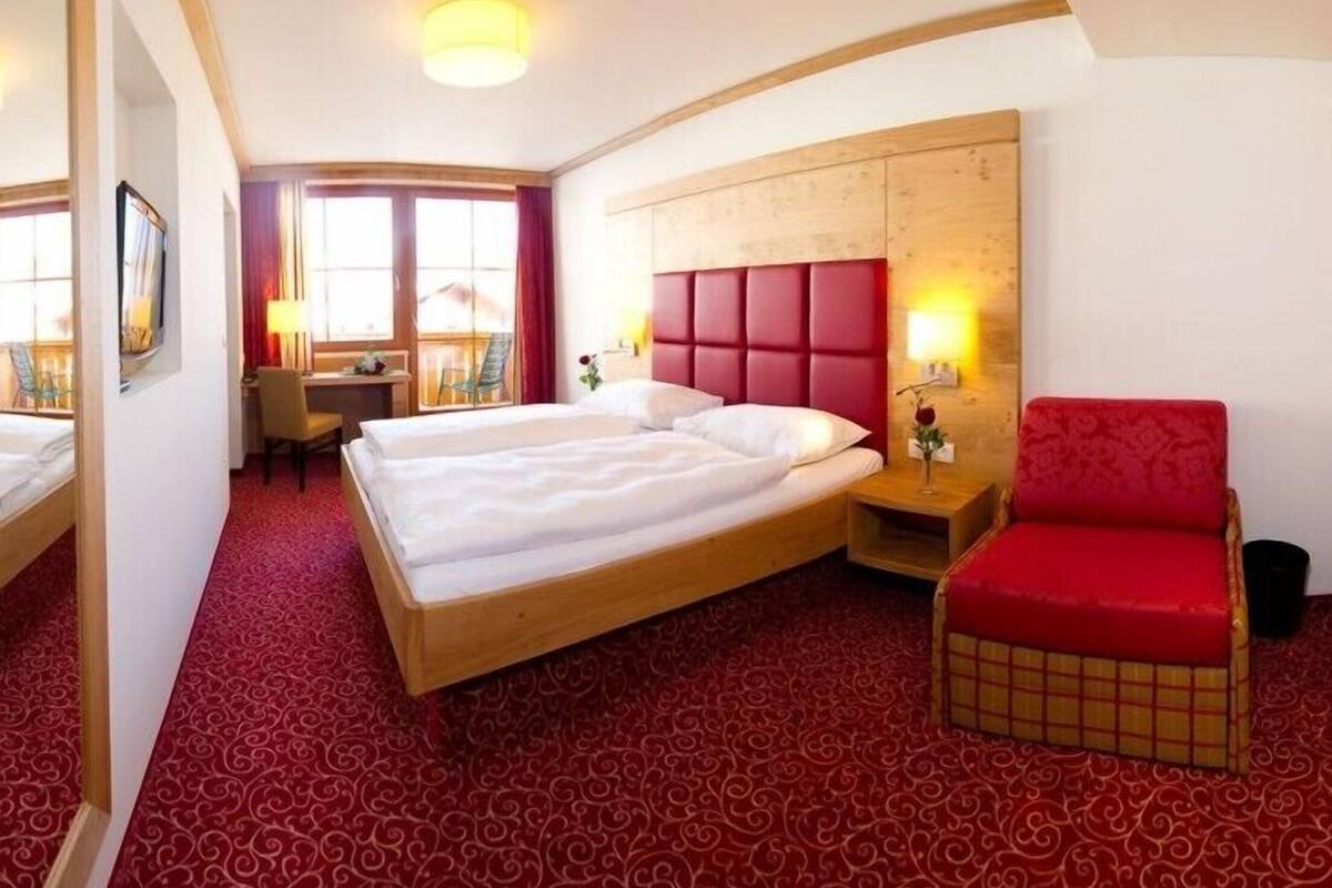A hotel room with a large bed, a red upholstered chair, a desk with a chair, and a large mirror.