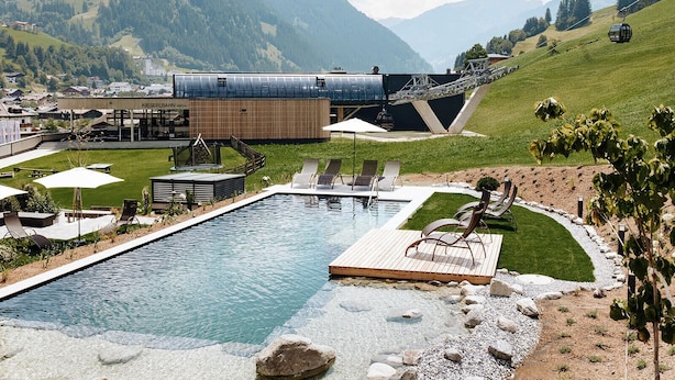 A swimming pool with lounge chairs, a wooden deck, and a view of mountains. - Hotel Roslehen (Grossarl)