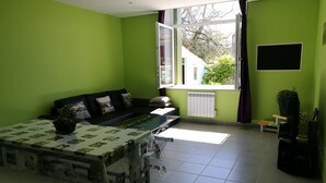 Ein Wohnzimmer mit grüner Wand, einem schwarzen Sofa, einem Glastisch und einem Fenster mit Blick auf Bäume.