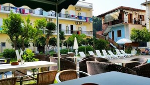 An outdoor seating area with wicker chairs and tables, a poolside lounge with sun loungers, and a building with balconies in the background. - Central Hotel (Parga)