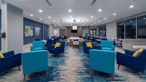Un moderno vestíbulo de hotel con sillones azules, una mesa de comedor central y una pantalla de televisión.