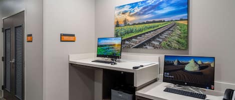 A hotel reception desk with two monitors, a keyboard, a mouse, and a printer. A sign indicates the business center is open 24 hours.