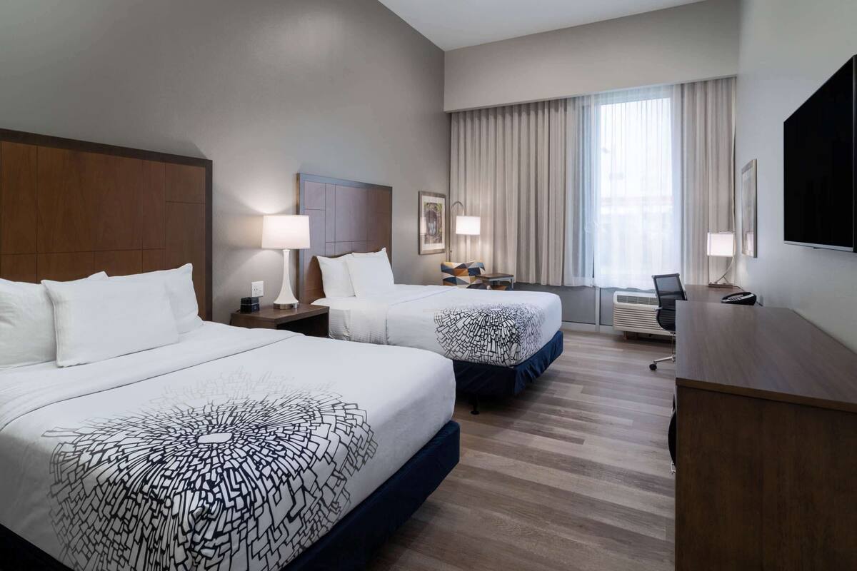 A hotel room with two beds, a desk, a chair, a TV, and a window with curtains.