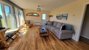A spacious living room with wooden flooring, a sofa, a coffee table, and large windows offering a view of the outdoors. - One-minute walk to a sandy beach. Beachside Haven Cottage awaits. (Belle River)