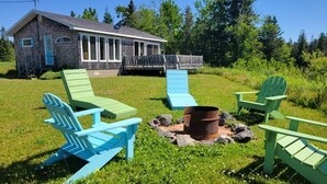 A rustic cabin with a deck, surrounded by green lawn chairs and a fire pit. - One-minute walk to a sandy beach. Beachside Haven Cottage awaits. (Belle River)