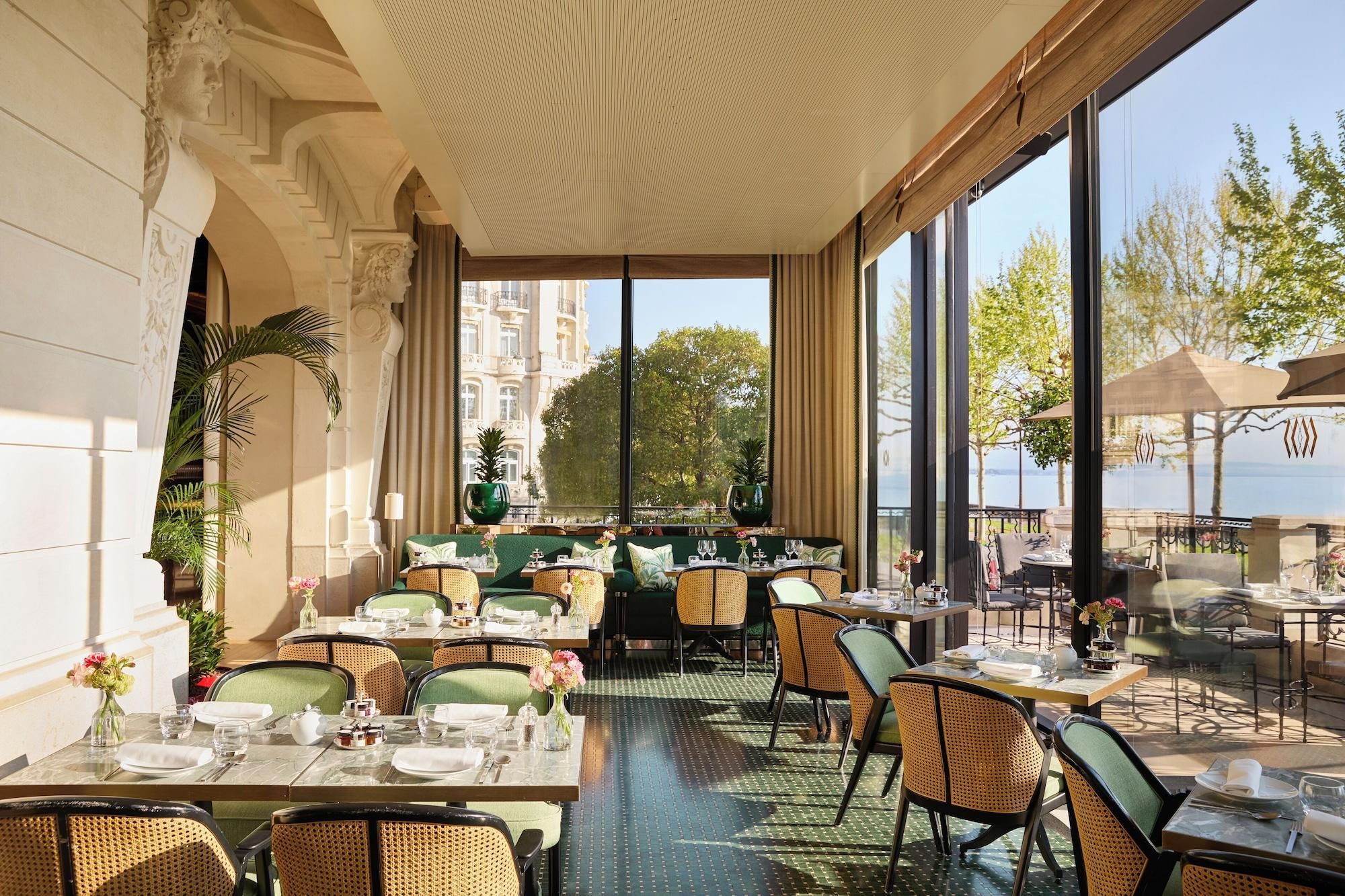A restaurant with outdoor seating, large windows, and a view of a building and trees.