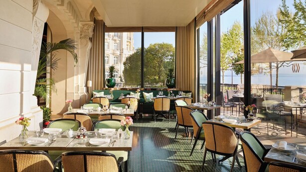 A restaurant with outdoor seating, large windows, and a view of a building and trees. - The Woodward An Oetker Collection Hotel (Geneva)